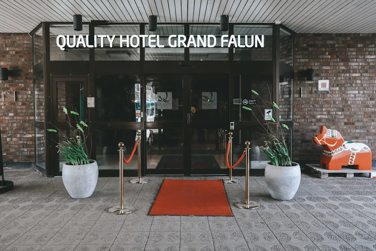 Glass entrance with red carpet leading to "QUALITY HOTEL GRAND FALUN." Flanked by potted plants; to the right, a colorful Dala horse statue stands against a brick wall.