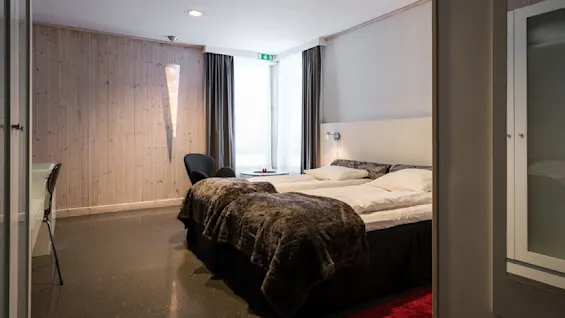 En pænt redt dobbeltseng med bløde tæpper står i et hyggeligt hotelværelse med trævægge, en stol ved et vindue og en skrivebordsstol. En lille loftslampe lyser blødt. Et nødudgangsskilt øverst viser "EXIT."