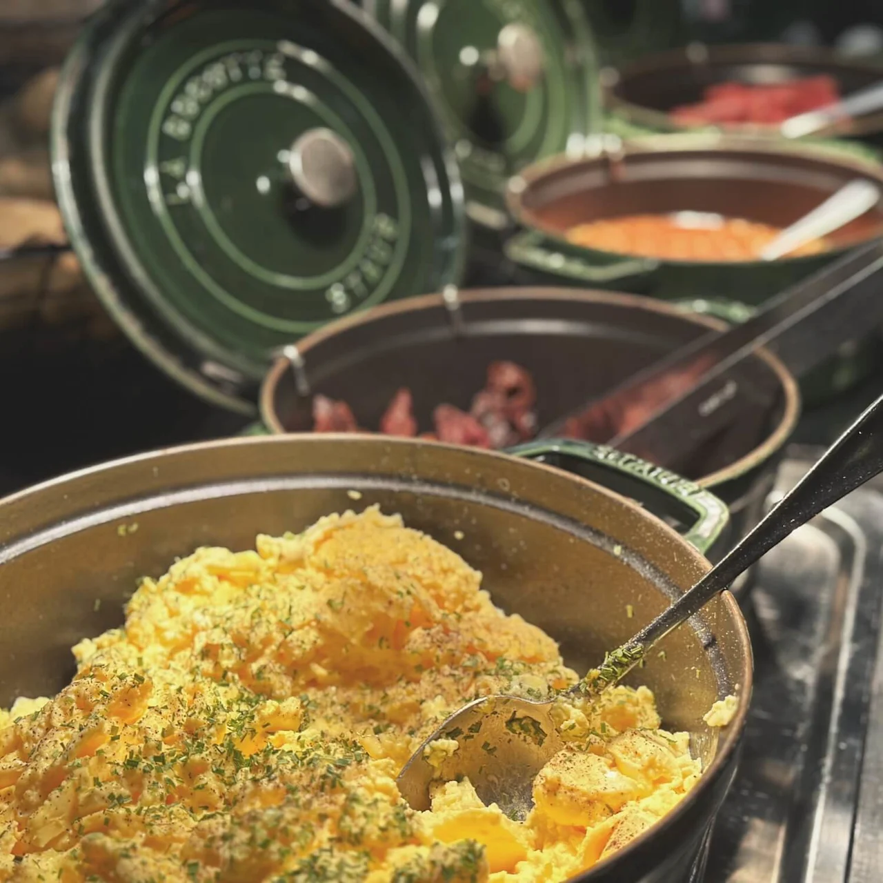 Scrambled eggs sit in a pan with a spoon, garnished with herbs. Background displays pots containing various dishes in a breakfast buffet setting.