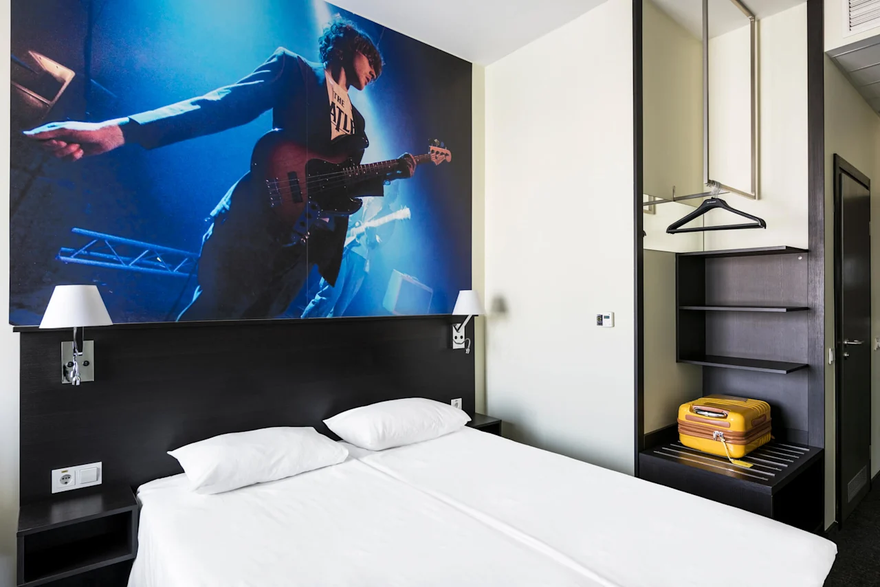 A hotel room features a double bed beneath a wall mural of a guitarist on stage. A wardrobe with shelves and a yellow suitcase is on the right.