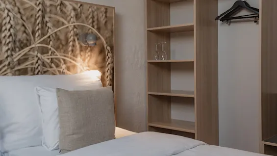 Viihtyisä hotellihuone: sänky, vehnäkuvioitu sängynpääty, yöpöytälamppu ja avoin vaatekaappi hyllyineen ja henkareineen.