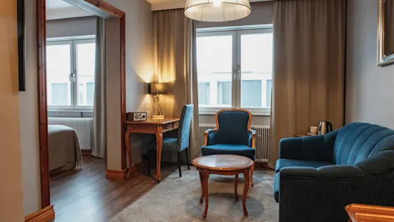En mysig hotellsvit med en vardagsrumsdel med blå sittplatser och ett skrivbord, som leder in till ett sovrum med en bekväm säng.