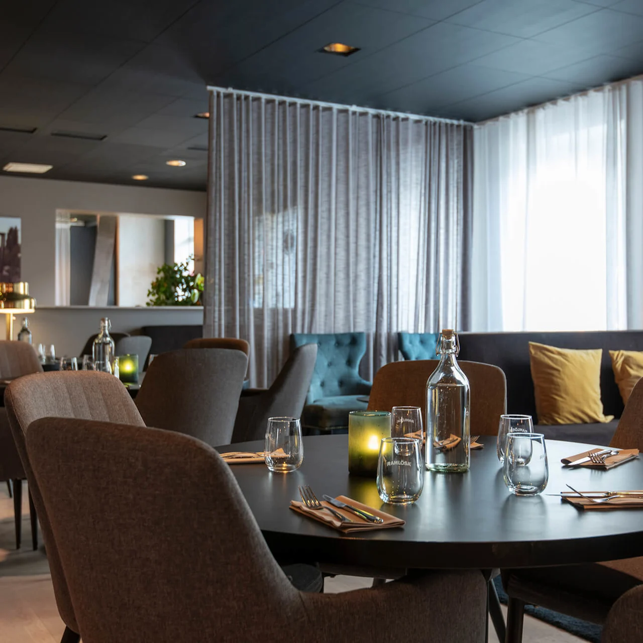 Ett runt bord är dukat med glas och bestick i en mysig restaurang, omgivet av bekväma stolar och mjuk belysning. Stora fönster med tunna gardiner lyser upp utrymmet.