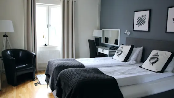 Bright hotel room featuring twin beds, a dark armchair by the window, and a small work desk.