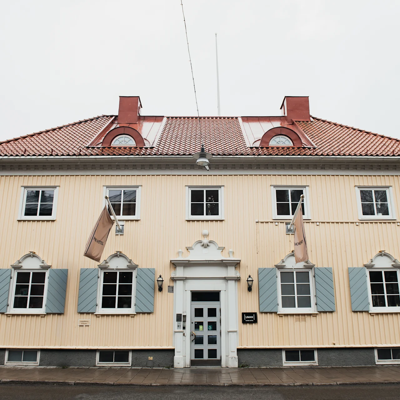 A beige wooden building with a red tiled roof, features six windows with blue shutters and three flags, standing on a paved street. 
