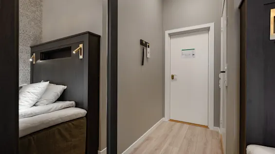 En hotelværelsesindgang med en seng til venstre, en lukket dør lige frem og en anden dør til højre. Sengen har læselamper.