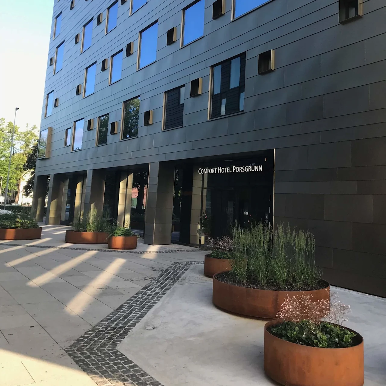 En moderne hotelfacade med reflekterende vinduer. Store plantekasser med grønt står på det mønstrede fortov. Skiltet lyder "COMFORT HOTEL PORSGRUNN".