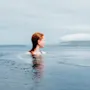 En person med rött hår i en infinitypool, som blickar ut över ett lugnt havslandskap med avlägsna öar under en molnig himmel.