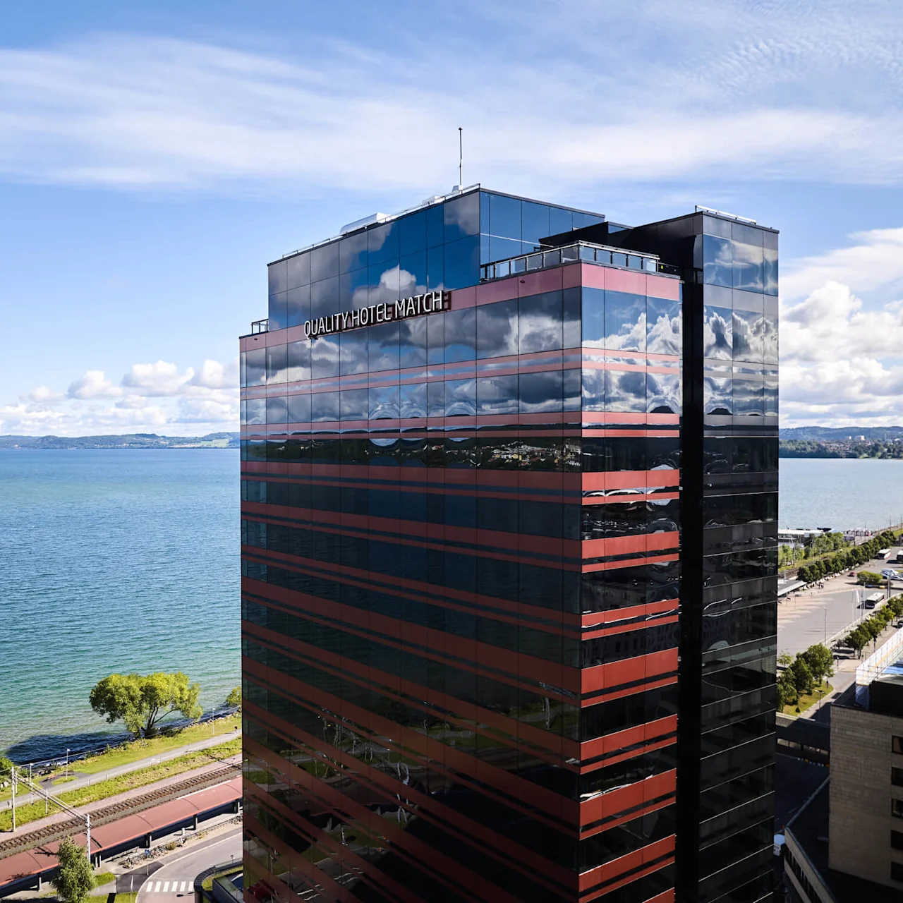 En høj bygning mærket "QUALITY HOTEL MATCH" står ved siden af en stor, rolig sø under en klar blå himmel, med fjerne bjerge og en træbevokset strand i baggrunden.