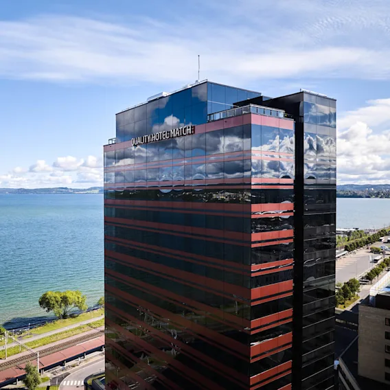 En høy bygning merket "QUALITY HOTEL MATCH" står ved siden av en stor, rolig innsjø under en klar blå himmel, med fjerne fjell og en tre-kantet strand i bakgrunnen.