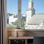 En skrivebordslampe hviler på et trebord foran et vindue med gardiner, med udsigt over en bysilhuet, der inkluderer et kirketårn under en blå himmel.