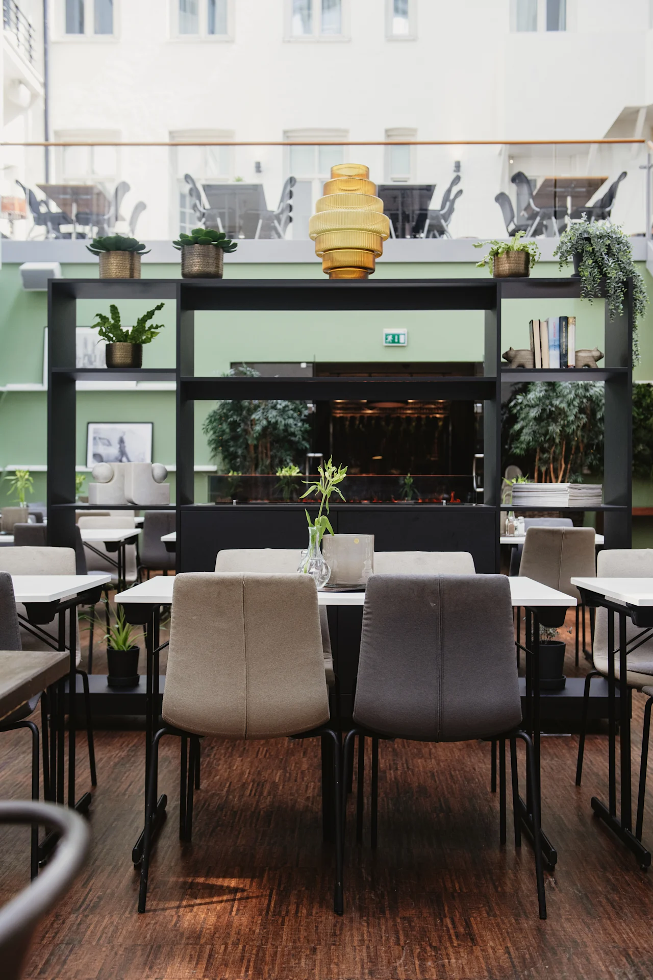 Stolar och bord är arrangerade i ett modernt, inomhus café med gröna väggar. Krukväxter och böcker dekorerar hyllor, medan naturligt ljus strömmar in genom stora fönster ovanför.
