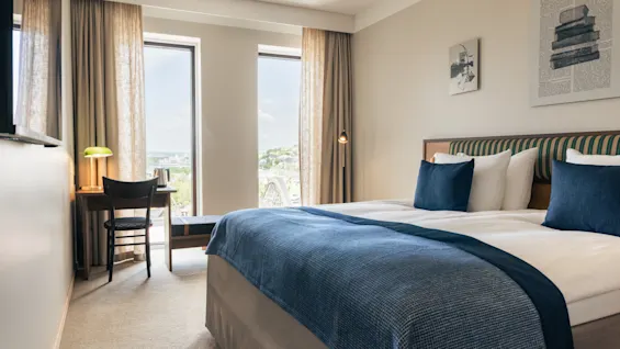 Et lyst hotellrom med en komfortabel seng, et skrivebord ved vinduet og utsikt over byen.