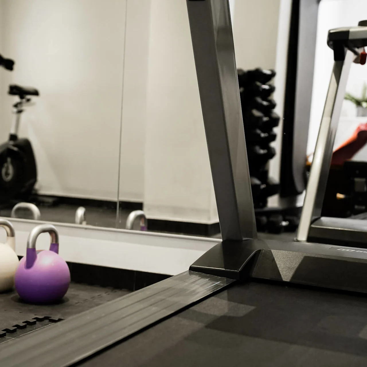 Kettlebells som ligger på gymmattor, omgivna av ett löpband, motionscykel och spegelvägg i ett träningsrum.