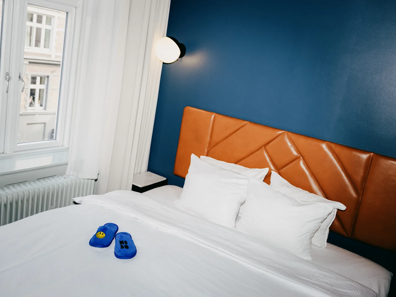 Moderni hotellihuone, jossa on sininen seinä, nahkainen sängynpääty ja siniset tossut sängyllä.