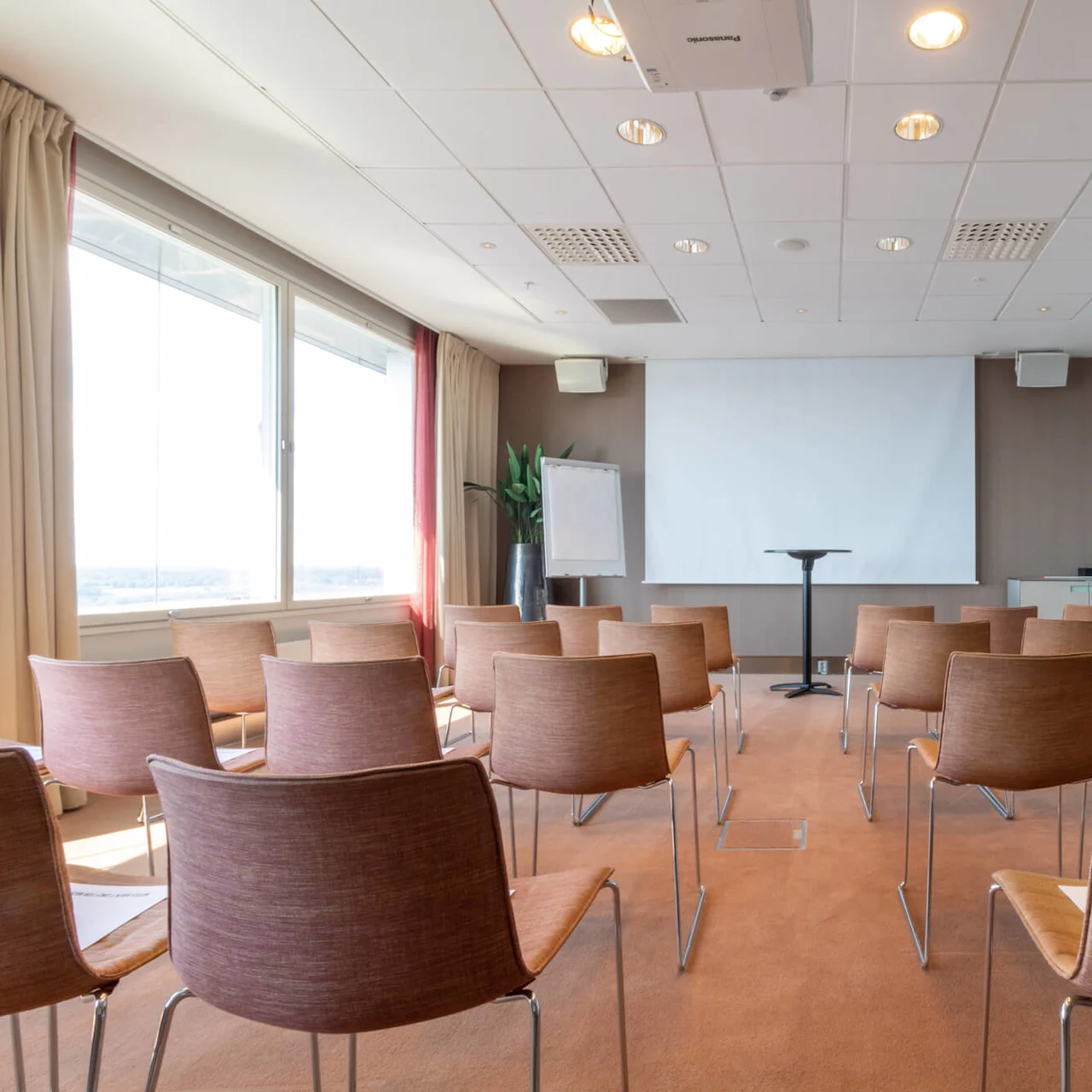A conference room with rows of brown chairs facing a blank projection screen on the wall. A flip chart is beside the screen, and large windows line the side.