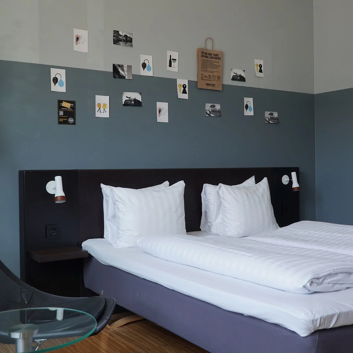 A neatly made double bed in a modern bedroom with pillows aligned, surrounded by a dark chair and round glass table. Wall displays are decorated with small art pieces.