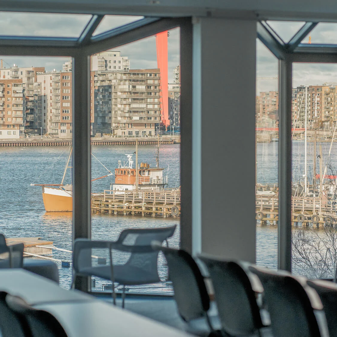 Office chairs are positioned around tables, viewed through large windows overlooking a harbor with docked boats and distant apartment buildings under a cloudy sky.