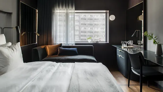 En pent senger står i et moderne hotellrom med tregulv. En sofa står nær et vindu med gjennomskinnelige gardiner og et skrivebord med glass og lampe.