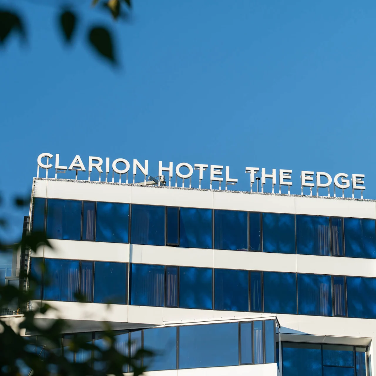 "CLARION HOTEL THE EDGE"-skiltet oven på en moderne bygning med blåtonede vinduer, set nedefra, omgivet af blade mod en klar blå himmel.