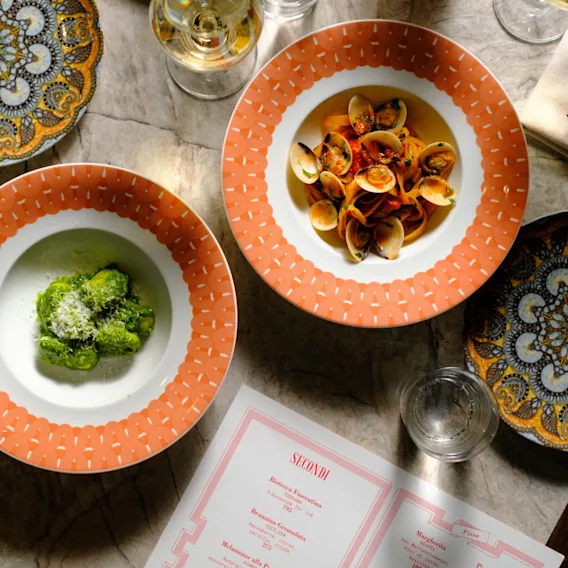 Dining table with two pasta dishes, one green, one with clams. Decorative plates, drinks, and a menu showing SECONDI are set.