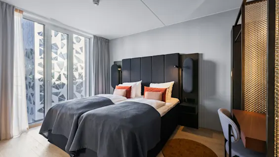 A neatly made double bed with gray blankets and orange cushions stands against a paneled wall. Sunlight filters through large windows with geometric patterns, illuminating a modern, minimalist room.