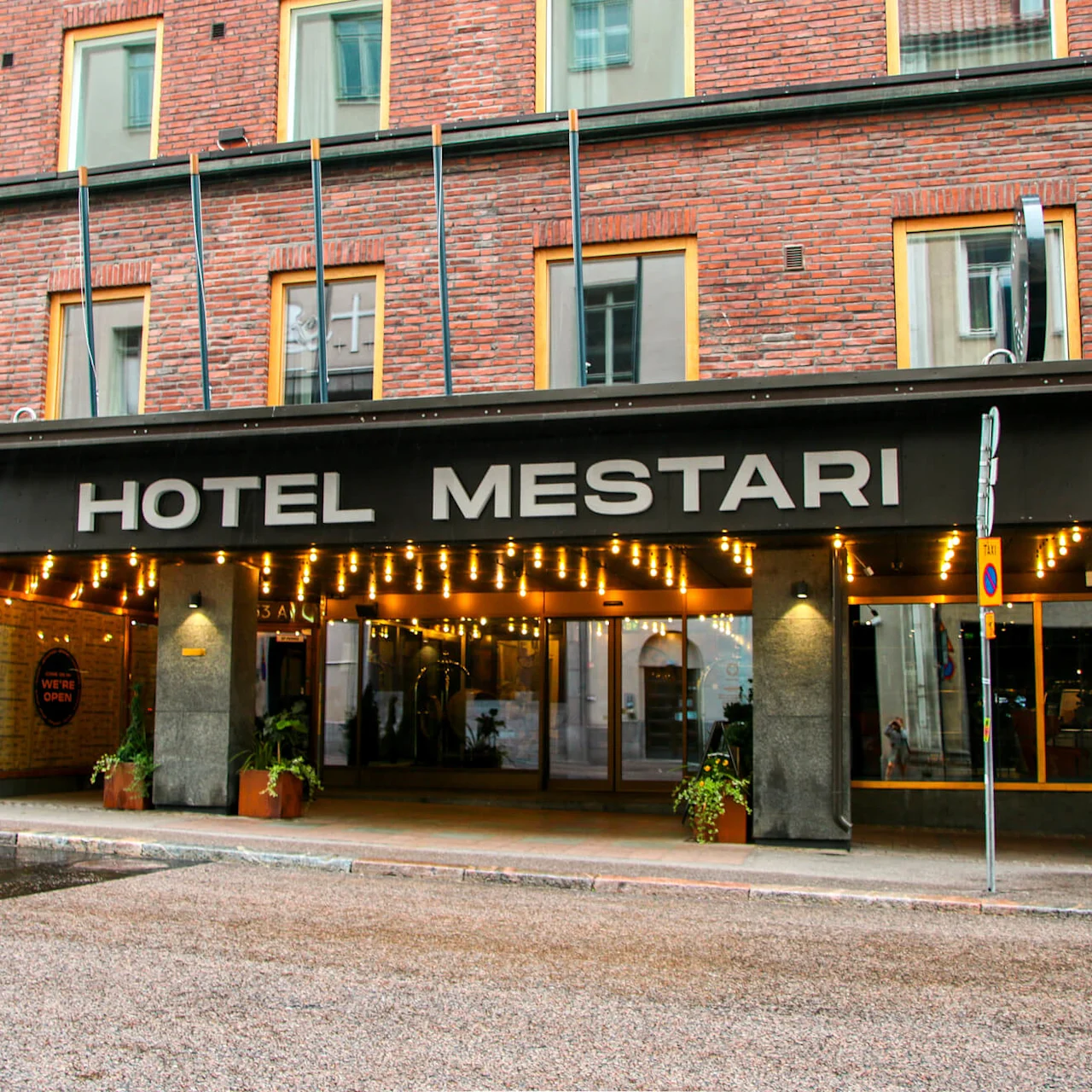 En hotellinngang viser navnet "HOTEL MESTARI" under lysende lys. Murbygningen har store vinduer. Et skilt lyder "WE’RE OPEN" med planter ved inngangen.
