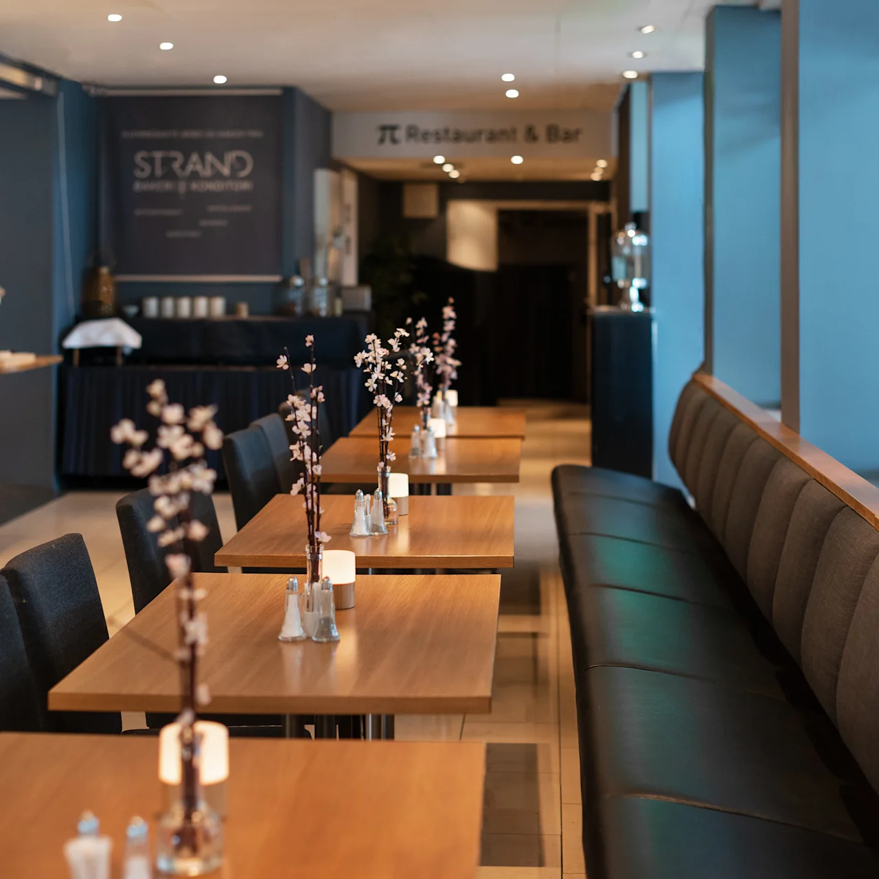 Bord med blomsterarrangemang kantar en restaurang med vadderade sittplatser och modern inredning. Skyltar visar "Strand" och "Restaurant & Bar."
