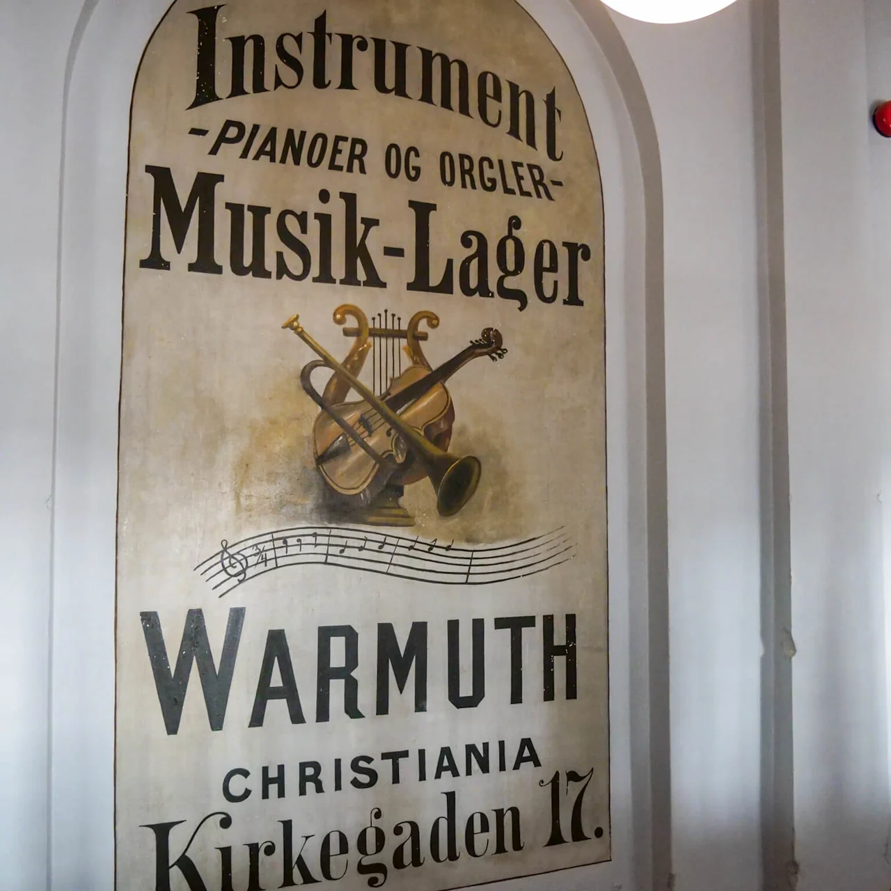 An old advertisement displays musical instruments like a trumpet and violin above text: "Instrument -Pianoer og Orgler- Musik-Lager Warmuth Christiania Kirkegaden 17." It’s painted on a wall. 