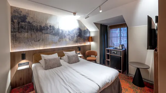A neatly made double bed is centered in a hotel room, surrounded by a modern desk, chair, and abstract art on the wall, with a window brightening the space.
