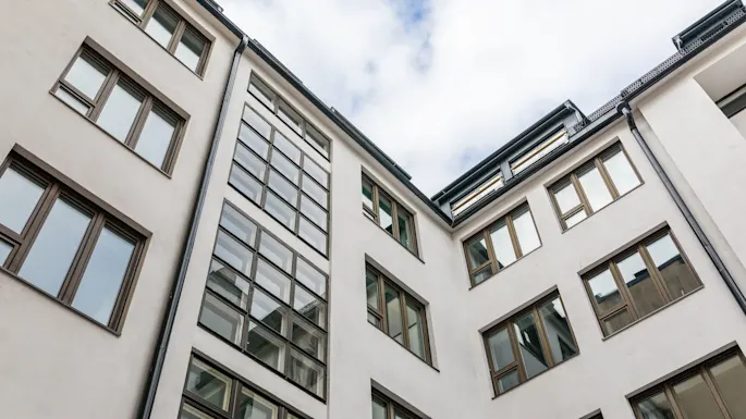 Et fleretagers bygning med store, reflekterende vinduer, har hvide pudsede vægge. Bygningen ses nedefra mod en skyet himmel.