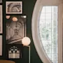 Flera inramade svartvita arkitekturfotografier hänger på en mörk vägg, upplysta av en sfärisk lampa; ett ovalt fönster med rutnät skymtar grönska utanför.