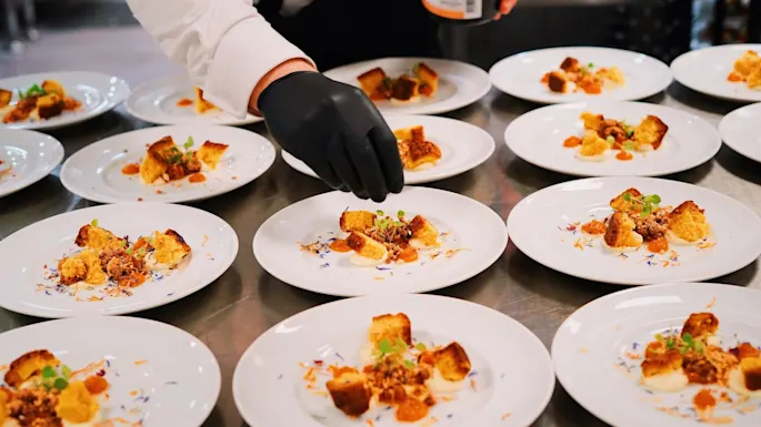 A chef with black gloves decorates multiple white plates with gourmet dessert toppings on a stainless steel kitchen counter.