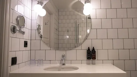 En rund spegel reflekterar ett kaklat badrum med starka lampor. En vit bänkskiva har en kran och två flaskor. Kontexten inkluderar en väggmonterad förstoringsspegel och två glas.