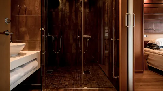 A modern bathroom with a glass shower enclosure and rusty-brown tiles, next to a bedroom with a visible bed.