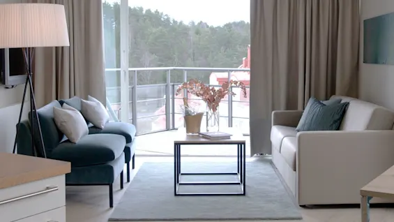 En moderne stue med en beige sofa og en blå lenestol rundt et salongbord med tørkede blomster på et teppe. Store vinduer viser en balkongutsikt mot skogen.
