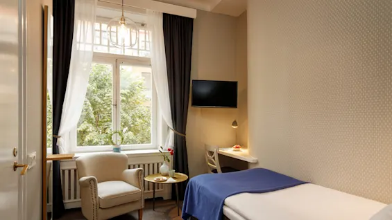 Ett mysigt hotellrum med en enkelsäng täckt med en blå filt, en fåtölj och ett litet bord. Ett fönster med tunna gardiner vetter mot grönska.
