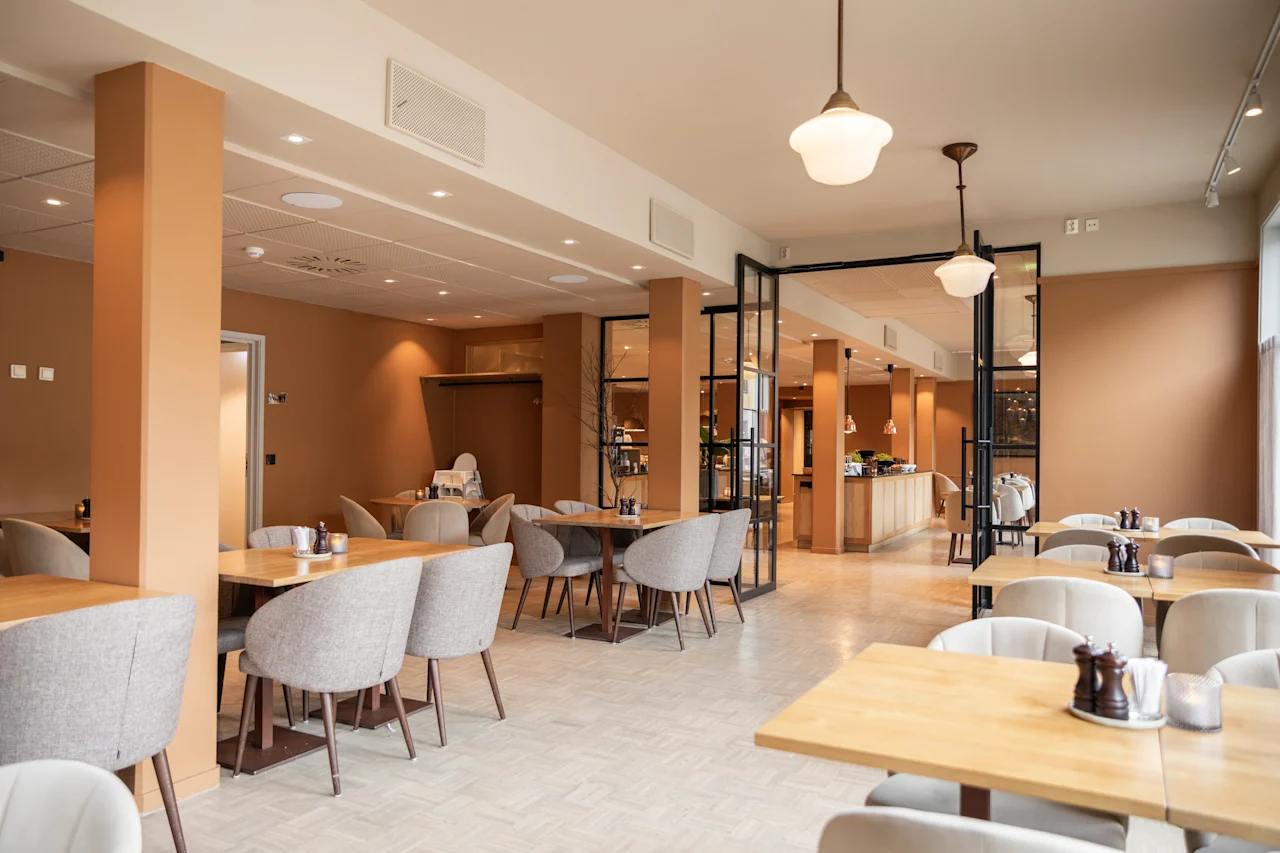 En restaurant med beige stoler og trebord viser et tomt sitteområde. Moderne lysarmaturer henger fra taket. Glassvegger deler opp rom, og skaper en innbydende atmosfære.