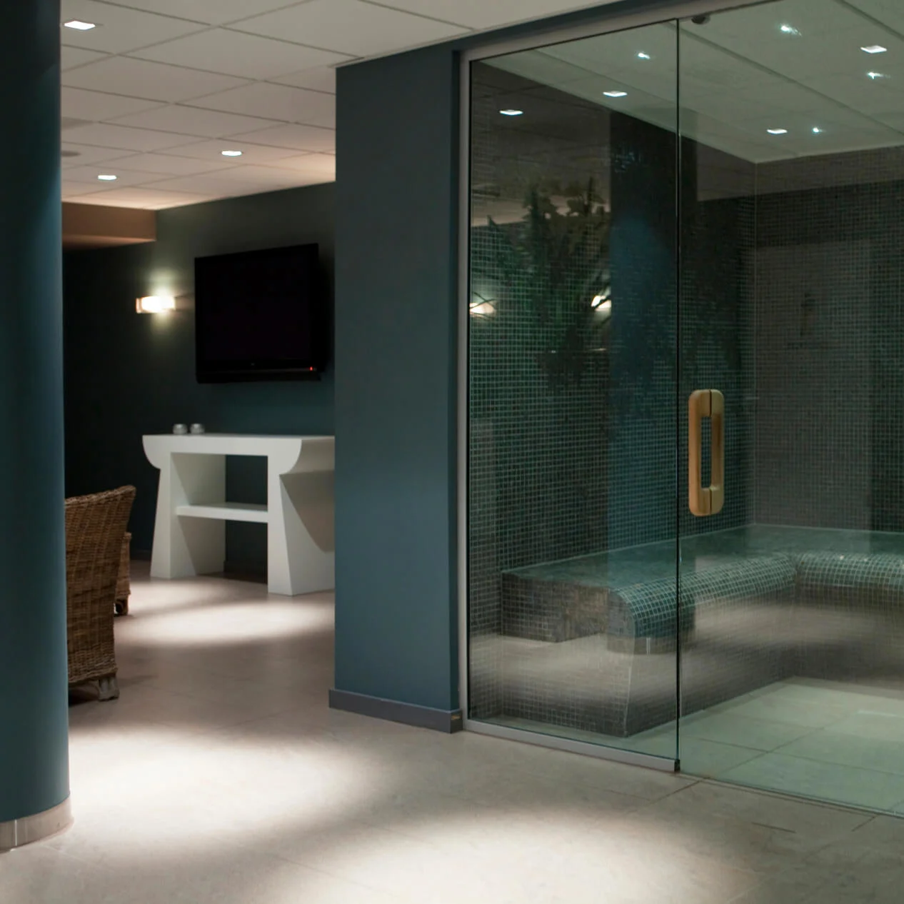 Glasindkapslet dampbad med flisebænke, placeret i et moderne spa-miljø. Tilgrænsende område viser kurvestol, vægmonteret TV og et dekorativt konsolbord mod mørke vægge.