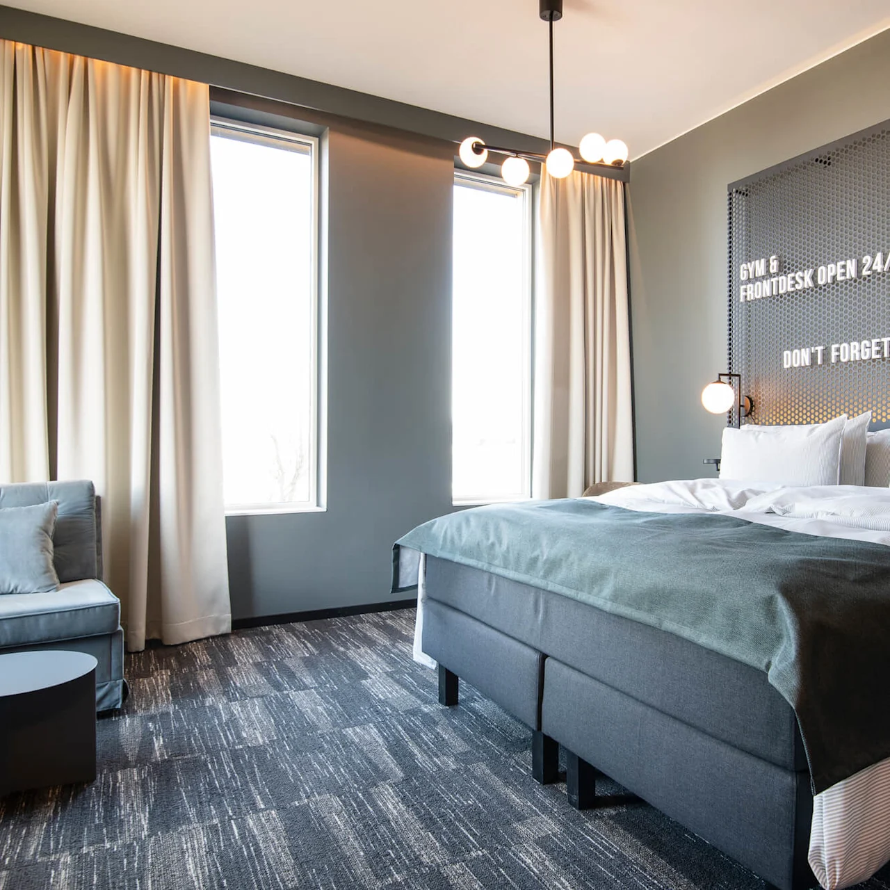 A neatly made bed with white sheets stands in a modern hotel room. White curtains and walls create a bright atmosphere. Text reads: "GYM & FRONTDESK OPEN 24/7. DON'T FORGET TO '#' THE BOX HOTEL."
