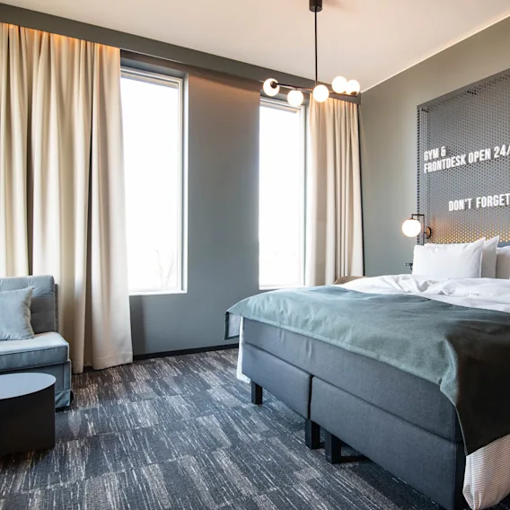 En prydligt bäddad säng med vita lakan står i ett modernt hotellrum. Vita gardiner och väggar skapar en ljus atmosfär. Texten lyder: "GYM & FRONTDESK OPEN 24/7. DON'T FORGET TO '#' THE BOX HOTEL."