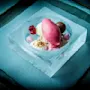 En farverig dessert med sorbet, mousse og bær arrangeret i en udhugget isbowle, placeret på en blå stofovertrukket overflade.