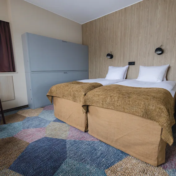Två prydligt bäddade enkelsängar med bruna filtar står i ett hotellrum. En mönstrad, färgglad matta täcker golvet, medan ett fönster med stängda gardiner och en stoppad stol fulländar miljön.