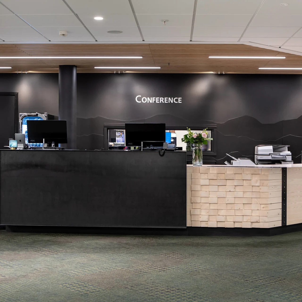 Reception desk with computers and flower vase; background features a black wall with the text "CONFERENCE" in a modern office setting.