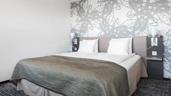 En prydligt bäddad säng med vita kuddar och en brun filt står i ett modernt hotellrum, prytt med abstrakta gråa väggmönster och sänglampor på varje sida.