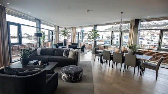 Luksuriøst stue med stor grå sofagruppe og spisebord, med utsikt over byen gjennom store vinduer. Rommet er pyntet med planter og en lysekrone.