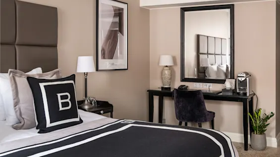 Seng med sort-hvidt sengetøj viser et "B", står ved siden af et mørkt skrivebord og stol med lampe og kaffemaskine. Beige vægge omgiver det moderne interiør.