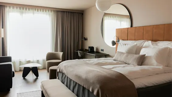 A cozy hotel room features a large bed, desk, and seating area. Sunlight streams through sheer curtains, creating a warm ambiance. Neutral colors enhance the modern, minimalist decor.