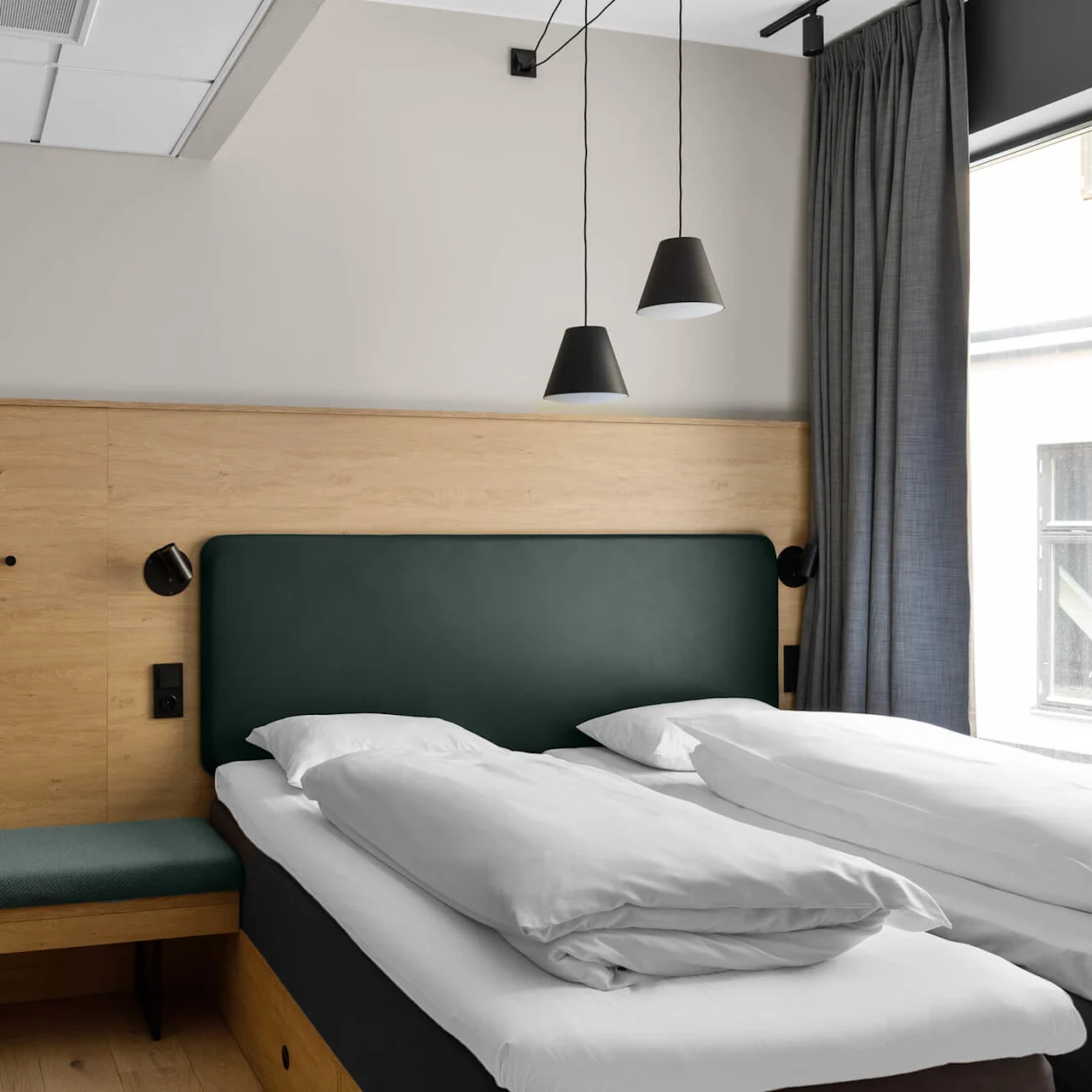 A modern hotel room with a wooden headboard features a double bed with white linens, a small upholstered bench, pendant lights, and a window with gray curtains.
