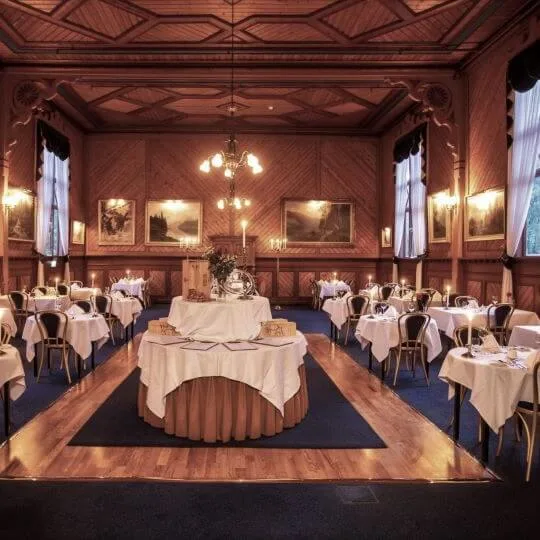 Elegant dining room with white tablecloth-covered tables arranged around a central display table. Wood-paneled walls, large windows with curtains, and chandeliers create a classic, upscale atmosphere.